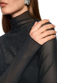 Frau, die ein transparentes schwarzes Oberteil mit kleinen funkelnden Details trägt, hält mit der rechten Hand ihre linke Schulter, sichtbares silbernes Ohrloch, ausgeschnittenes Gesicht.