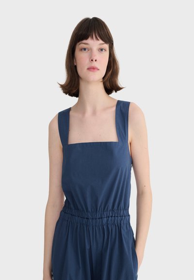 Femme aux cheveux bruns courts portant une combinaison sans manches bleu marine avec un décolleté carré et une taille élastique, debout devant un fond uni.