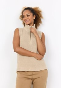 Beige ärmlös stickad polotröja med dragkedja fram, med ribbat mönster. Matchas med beige byxor, mot en vit bakgrund.