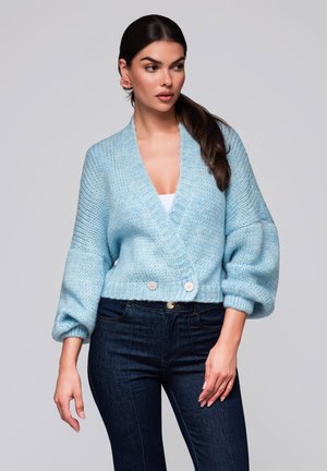 Lichtblauwe gebreide cardigan met een diepe V-hals, oversized mouwen en twee knopen bij de taille, gecombineerd met donkere spijkerbroeken.