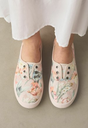 Pies con zapatos de lona florales sin cordones con suelas de goma blancas, combinados con una falda blanca, de pie sobre un suelo beige liso.