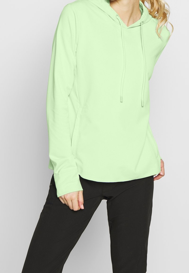 Femme portant un sweat à capuche vert clair à manches longues et un pantalon noir, debout devant un fond uni et clair.