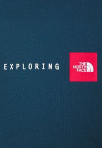 Tmavě tyrkysová tkanina s bílým textem "EXPLORING" a červeným čtvercovým logem s nápisem "THE NORTH FACE" bílými písmeny.