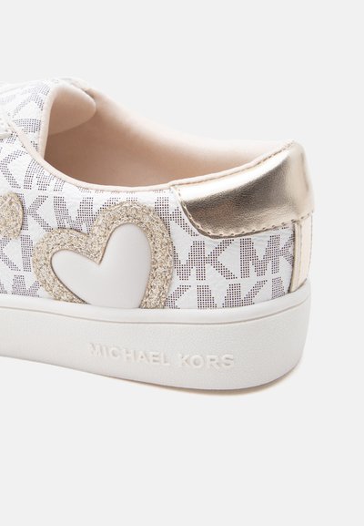 Άσπρο αθλητικό παπούτσι με μοτίβο λογότυπου MK σε χρυσό, λεπτομέρεια με λαμπερή καρδιά και μεταλλική χρυσή ταμπέλα στη φτέρνα πάνω σε λευκή λαβή από καουτσούκ με ανάγλυφο το όνομα της μάρκας.