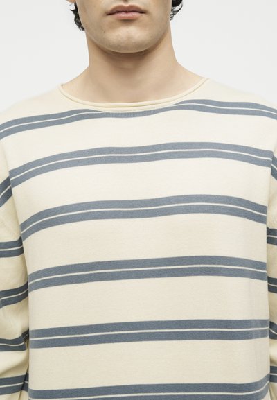 Homme portant un pull crème à rayures horizontales bleues, visible du cou jusqu'au milieu du torse sur un fond uni.