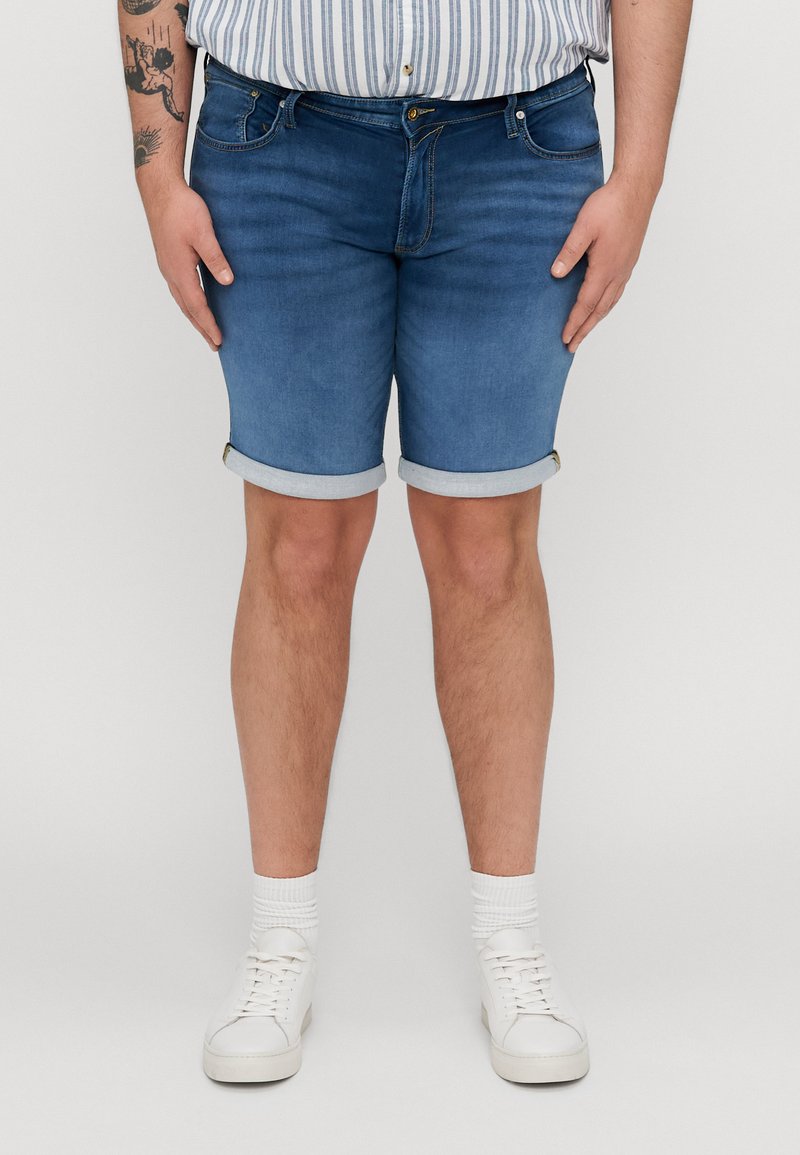 Shorts en denim bleu clair avec un ourlet roulé, présentant un style classique à cinq poches et une fermeture éclair. Assortis à des baskets blanches et des chaussettes.