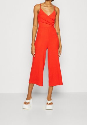 Mono rojo de canalé con un cuerpo ajustado, tirantes finos y piernas anchas y recortadas. Llevado con sandalias de tacón blancas.