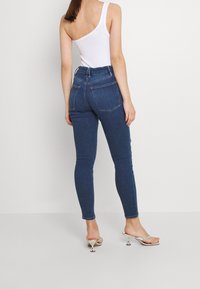 Vaqueros oscuros ajustados de mezclilla, con cintura alta y bolsillos traseros. Combinados con una blusa blanca de un solo hombro y sandalias plateadas de tacón.