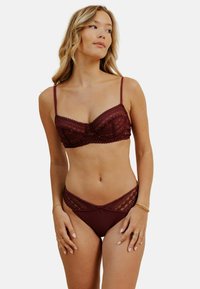 Bralette en dentelle bourgogne avec bretelles réglables, présentant des motifs géométriques. Bas de bikini taille haute en dentelle assortis. Texture lisse.