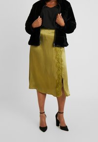 Chaqueta de piel sintética negra, blusa negra y falda de satén verde oliva asimétrica con detalle de encaje y abertura lateral. Zapatos de tacón alto negros.