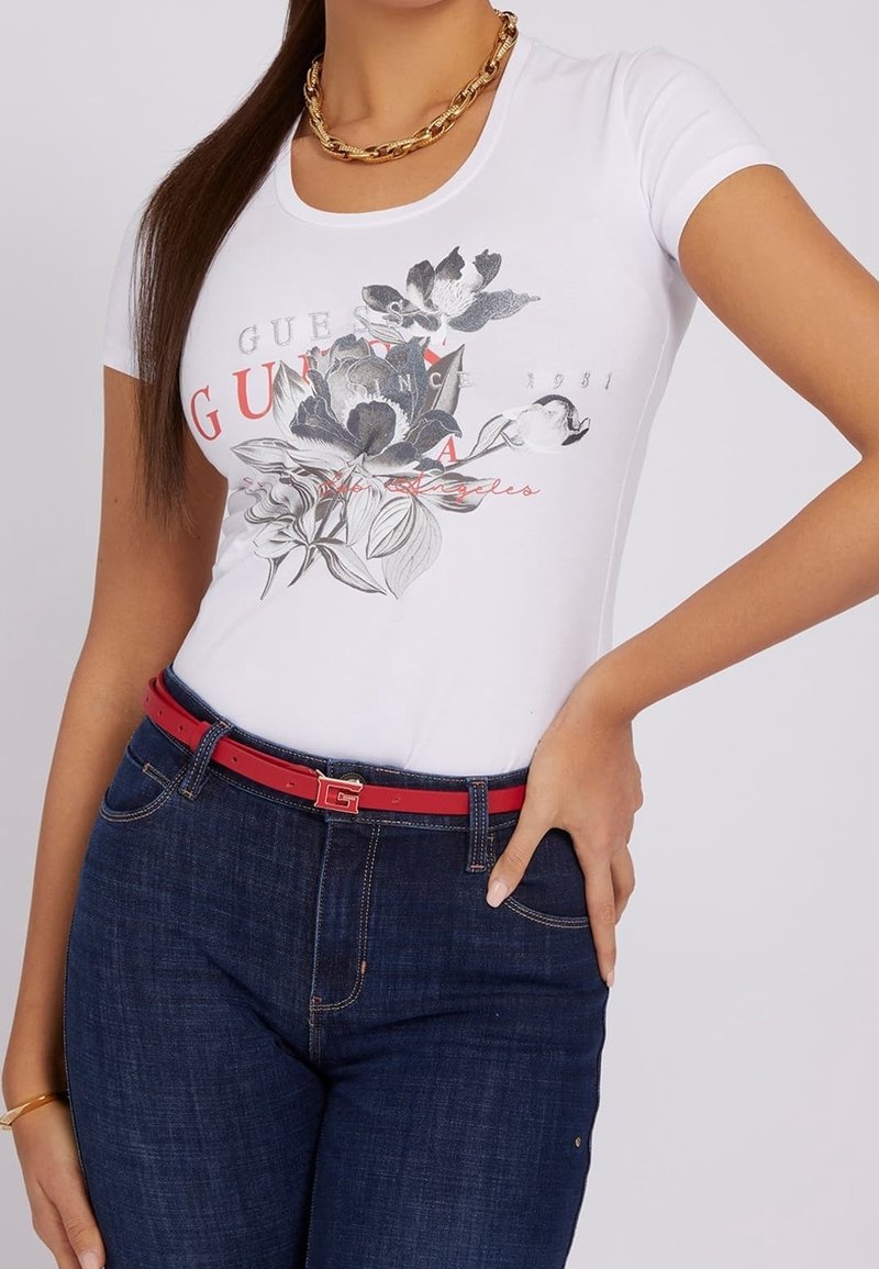 Femme portant un t-shirt blanc à imprimé floral, un jean bleu foncé, une ceinture rouge avec une boucle dorée, un collier chaîne dorée et un bracelet doré.