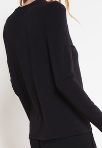 Černé tričko s dlouhým rukávem a kulatým výstřihem, měkká texturka látky a přiléhavý střih. Bezešrá záda a minimální detaily švů.