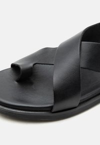 Μαύρο δερμάτινο σανδάλι με φαρδιές λουρίδες, που διαθέτει θηλιά για τα δάχτυλα και επίπεδη σόλα, παρουσιασμένο σε κοντινή λήψη.