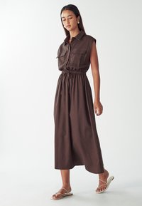 Hnedé šaty bez rukávov s golierom a dvoma prednými vreckami, s pásom na stiahnutie a splývavou sukňou po členky.