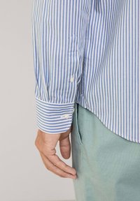 Lichtblauw en wit gestreept shirt met een knoopmanchet, dat een gladde katoenen textuur laat zien. Gecombineerd met groene broek, wat een casual look weergeeft.