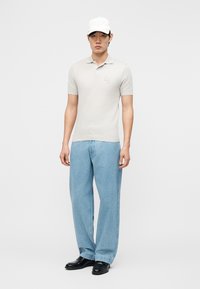 Hombre con gorra blanca, polo gris claro, vaqueros sueltos azul claro y zapatos negros, de pie contra un fondo blanco.