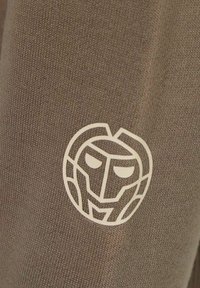 Un tessuto marrone presenta un emblema bianco che somiglia a un volto di leone stilizzato, caratterizzato da dettagli angolari e affilati e un contorno circolare.