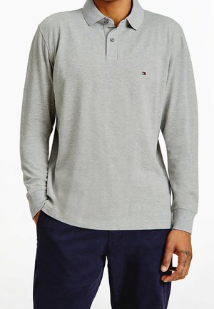 Homme portant un polo gris clair à manches longues avec col boutonné et pantalon foncé, main reposant près de la poche.