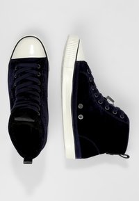 Pari siniset samettiset korkeakärkiset tennarit, joissa on valkoiset kumikärjet ja -pohjat, mustat nauhat ja metalliset silmukat valkoista taustaa vasten.