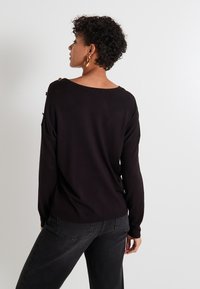 Un suéter negro con escote barco, mangas largas y detalles de botones dorados en el hombro. Tejido suave con un corte relajado. Combinado con jeans oscuros.