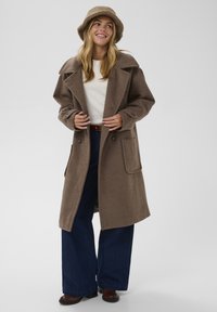 Brązowy, wełniany, luźny płaszcz z dużymi klapami i przednimi kieszeniami, noszony na kremowym swetrze, w połączeniu z rozszerzanymi dżinsami i kapeluszem typu bucket.