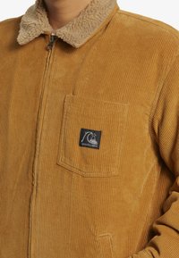 Kordfløjlsjakke i varm brun med en pelsagtig beige krave, har lodrette ribbet tekstur, frontlomme og logo-patch.
