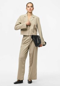 Beige cropped blazer med lange ærmer og en enkelt knap, parret med matchende bukser. Sort tekstureret clutch-taske og sorte flade sko.