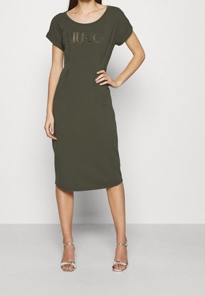 Mujer vistiendo un vestido verde oliva hasta la rodilla con mangas cortas y "LIU JO" impreso en el pecho, combinado con tacones plateados de punta abierta.