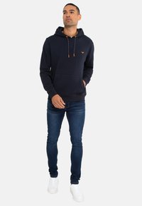 Uomo che indossa un hoodie blu navy con lacci arancioni, jeans skinny blu e sneakers bianche, in piedi con una mano in tasca.