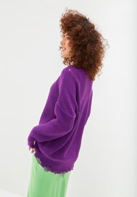 Maglione in maglia viola con un motivo testurizzato e orlo sfrangiato, abbinato a una gonna verde a coste, visto di lato su uno sfondo bianco.