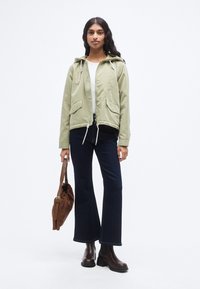 Χακί αδιάβροχο μπουφάν με κουκούλα, ρυθμιζόμενο στρίφωμα με κορδόνια και δύο εμπρόσθιες τσέπες, συνδυασμένο με σκουρόχρωμο καμπάνα τζιν και καφέ μποτάκια αστραγάλου.