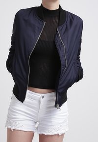 Chaqueta bomber marinera con puños acanalados, cierre de cremallera y bolsillos laterales, combinada con pantalones cortos blancos con dobladillo de crochet y una blusa negra texturizada.