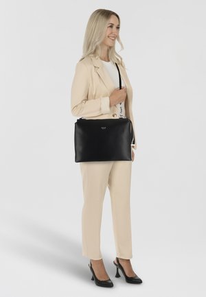 Zwarte leren handtas met ritssluiting, zilveren hardware en een afneembare schouderriem, vastgehouden door een persoon in een beige pak.