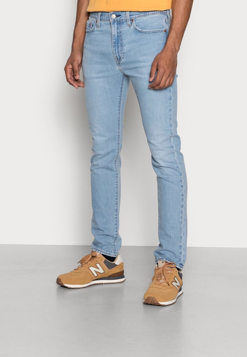 Ljust blå denimjeans med slim fit, parat med tan sneakers med vita accenter och en synlig logotyp, mot en neutral bakgrund.