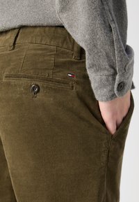 Pantalons en velours côtelé vert olive avec une finition texturée, présentant une poche avant, une fermeture à un bouton et un petit logo de la marque sur le côté.