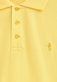 Gul polo skjorta i texturerad tyg. Har en tre-knapp placket och en broderad figur i nedre högra hörnet.