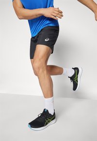 Pantalones cortos deportivos negros, camiseta de manga corta azul, zapatillas de correr negras con detalles verdes, calcetines blancos. Enfocado en el movimiento activo y la vestimenta deportiva.