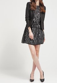 Černý blejzr přes vzorované šaty do kolen s vertikálními metalickými pruhy. Šaty mají přiléhavý pas a rozšířenou sukni. Černé lodičky.