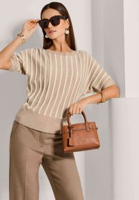 Vrouw in een beige gestreepte trui en broek, met grote oorbellen, een zonnebril, gouden armbanden, die een kleine bruine leren handtas vasthoudt en leunt tegen een witte muur.