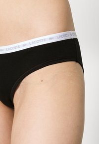 Juodos medvilnės žemos talijos bikini kelnaitės su balta elastinga juostele, ant kurios pilka spalva atspausdintas prekės ženklas "LACOSTE".