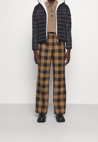 Veste noire matelassée avec doublure en polaire blanche, polo beige, et pantalon large à carreaux marron et noir. Bottes noires complètent la tenue.