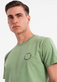 Camiseta de algodón verde con un gráfico negro redondo en el pecho, que presenta patrones moteados en toda la tela. Mangas cortas y cuello redondo.