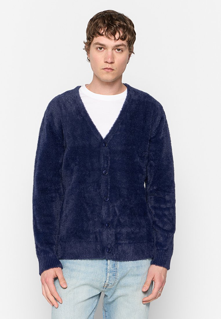 Jeune homme portant un cardigan boutonné en peluche bleu marine sur un T-shirt blanc et un jean bleu clair, se tenant devant un fond blanc uni.