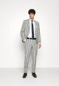 Abito a quadri grigio con blazer aderente e pantaloni slim. Indossato su una camicia bianca, completato da una cravatta nera e una cintura. Scarpe nere completano il look.