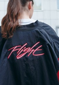 Černá bunda s volným střihem, na zadní straně s červeným vyšívaným nápisem "Flight", hladká textura a kulatý výstřih.