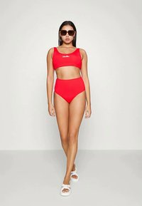 Ženska v rdečem bikiniju z visokim pasom in kratkim topom, s temnimi sončnimi očali in belimi sandali, stoji ob nevtralni svetli podlagi.