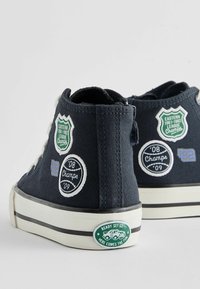Wysokie sneakersy z granatowego płótna z wieloma haftowanymi naszywkami oraz gumową podeszwą z białym paskiem. Zauważalne detale to naklejki akcentujące.
