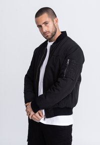 Giacca bomber nera con polsini a costine, chiusura frontale con cerniera e tasche laterali. Presenta una manica con motivo orizzontale a rilievo. Indossata sopra una camicia bianca.