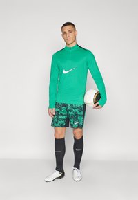 Grünes langärmliges Sportshirt mit schwarzen Akzenten, dazu passende gemusterte grüne Shorts, schwarze Socken und weiße Fußballschuhe, während er einen Ball hält.
