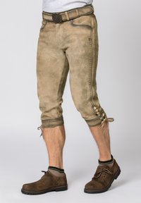 Braune Wildlederlederhosen mit kniehohem Schnitt, dekorativer Stickerei und Knopfakzenten an den Bündchen. Getragen mit braunen Schuhen und Socken mit grünen Akzenten.